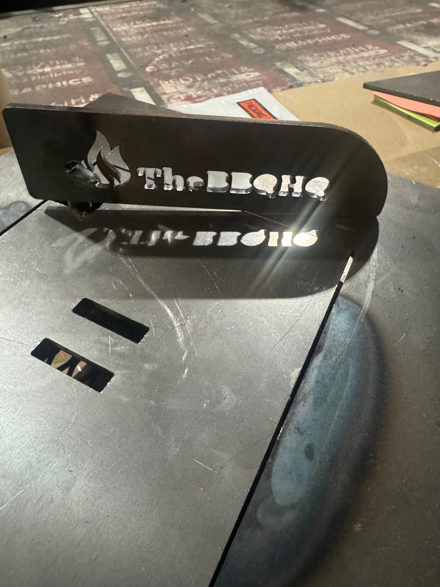 TheBBQHQ black metal grill sign with flame logo and white lettering on table
