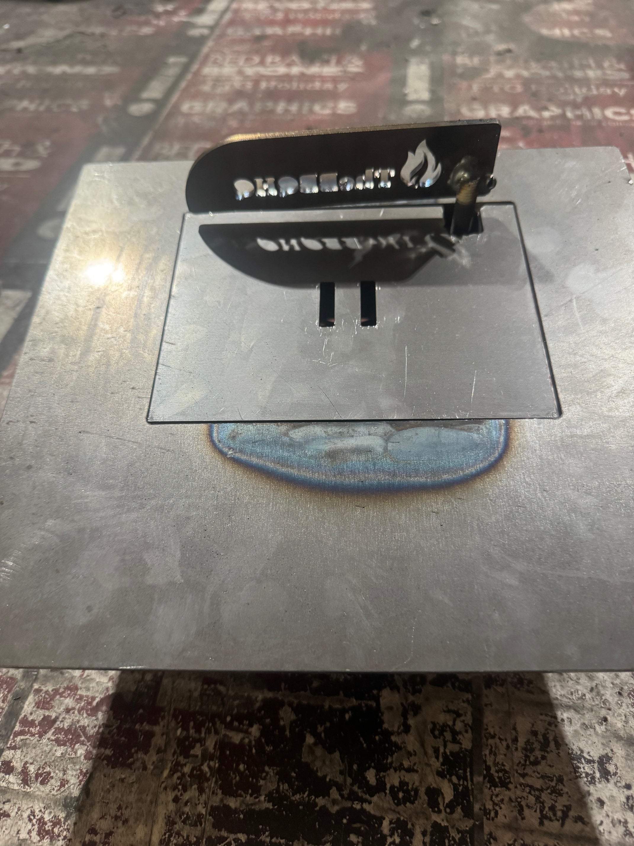 Black metal grill firebox with flame logo and text, mounted on stainless steel base plate