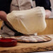Fresh pizza dough in stainless steel bowl on wooden surface with flour